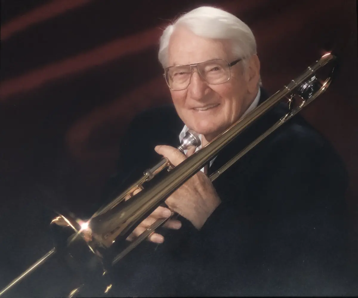 A portrait of an older man wearing glasses holding a trombone.